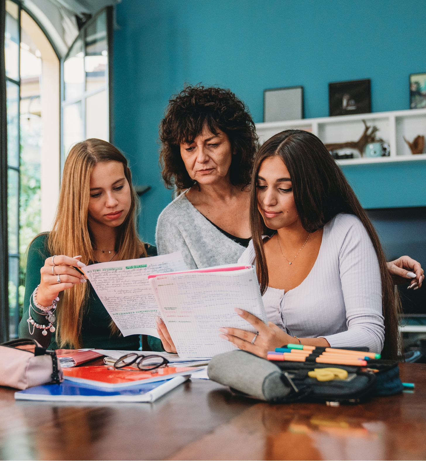A woman helping two teenage girls with their homework at a table.