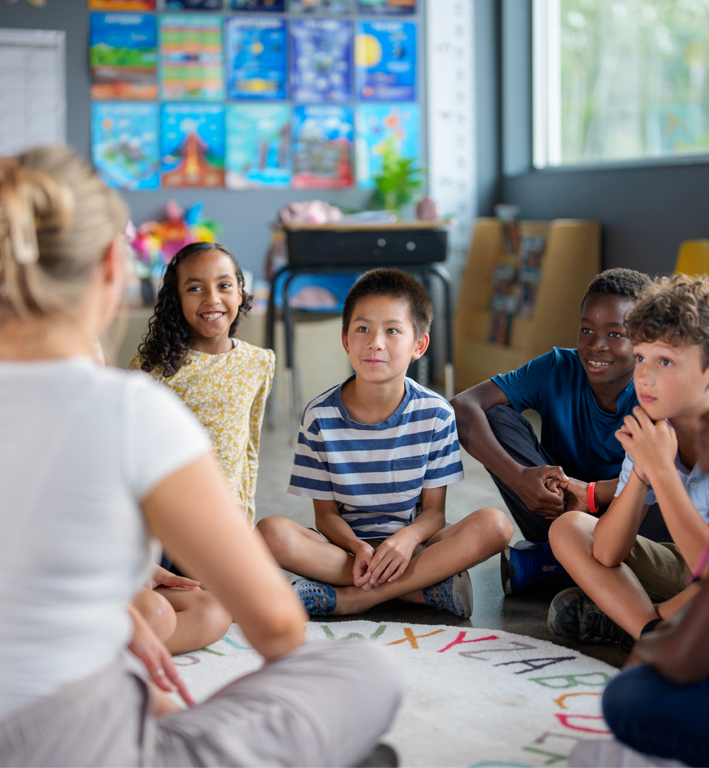 Children sitting on the floor listening to a teacher in a colorful classroom.